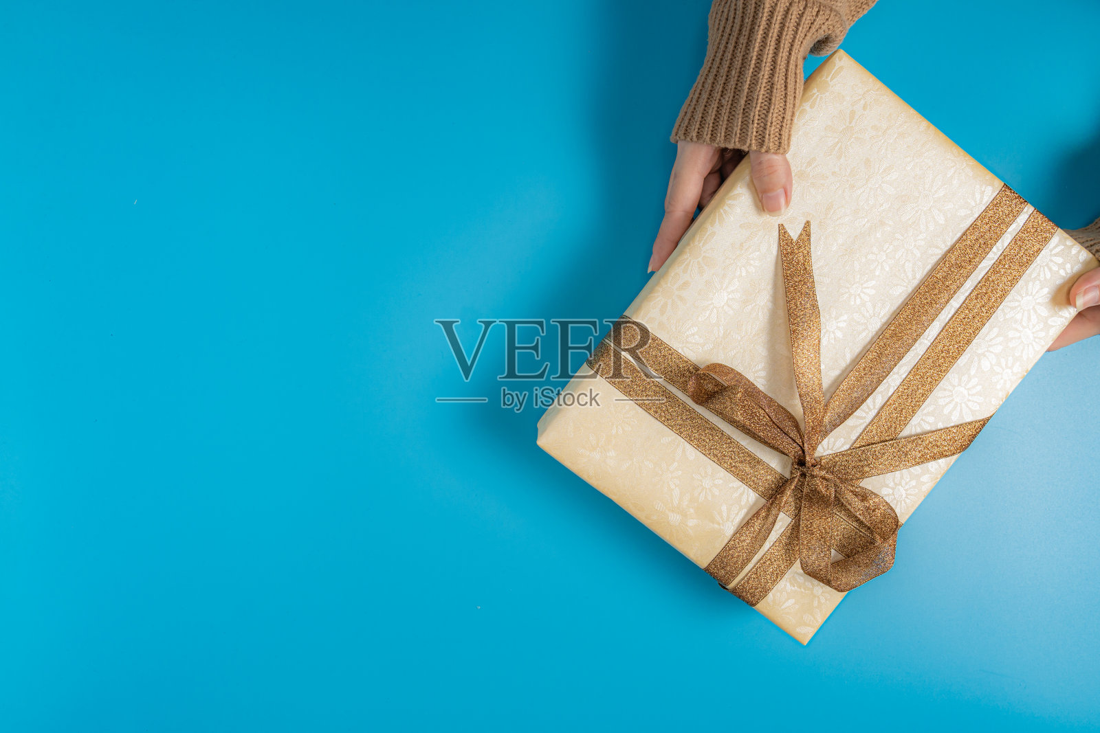 有人手穿一件棕色毛衣拿着一个礼品盒与棕色丝带系在蓝色背景位于右上角的图片照片摄影图片
