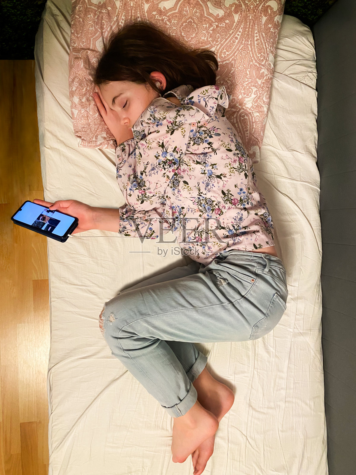 年轻疲惫的女孩在床上拿着智能手机侧身睡觉，真人从上面偷拍照片。照片摄影图片