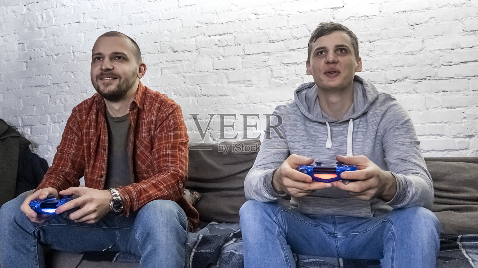 两个人在白砖墙前玩电子游戏。双手握着控制台控制器。男生坐在沙发上，正面照片摄影图片