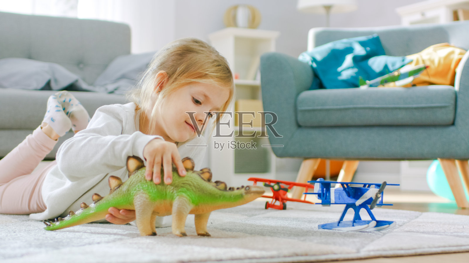 可爱的金发小女孩躺在家里的地毯上，玩玩具恐龙和飞机。快乐的孩子在阳光明媚的客厅玩玩具。特写肖像拍摄。照片摄影图片