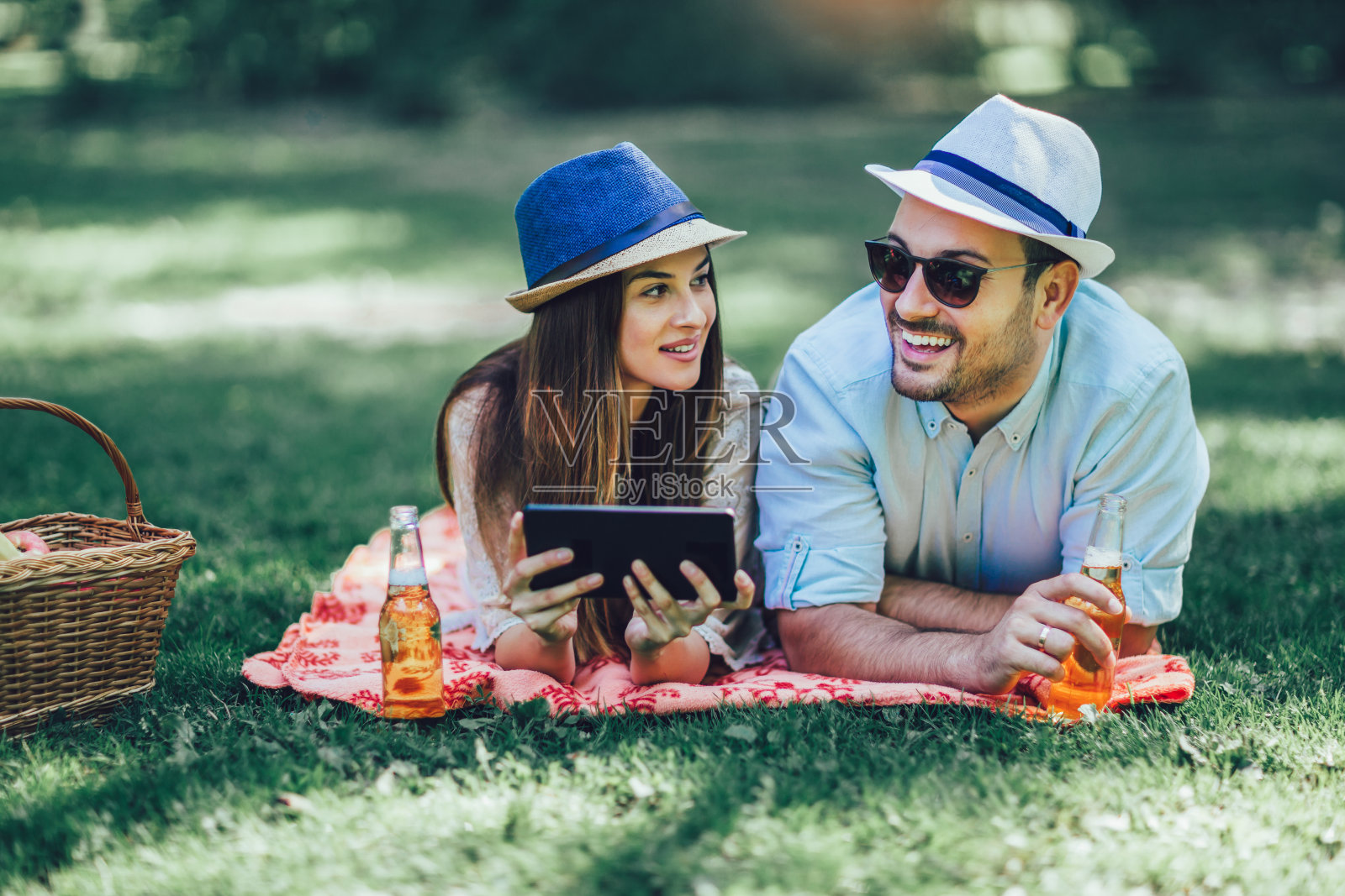 一对情侣躺在公园里的野餐毯子上，篮子里装满了水果，他们用的是数码平板电脑照片摄影图片