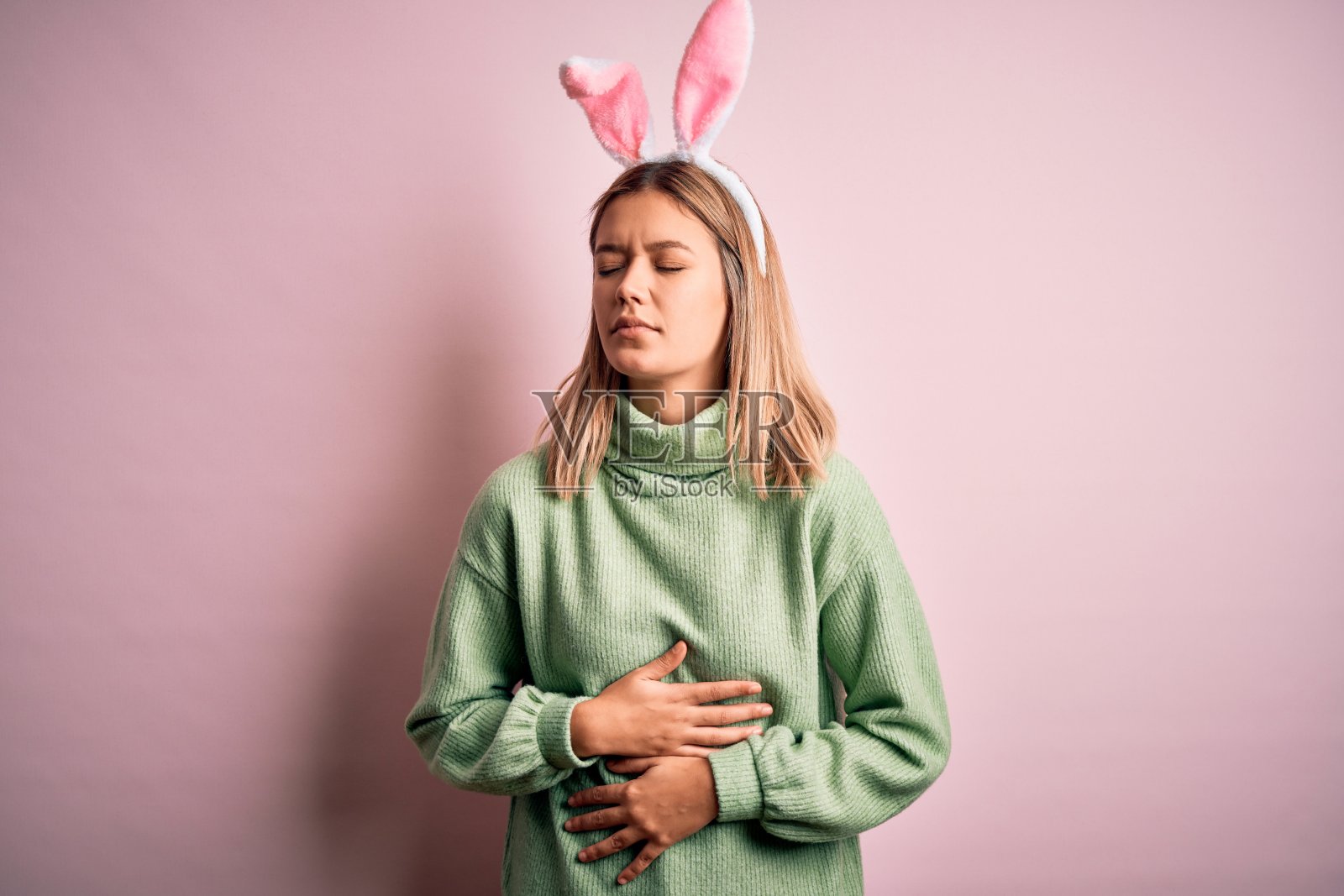 年轻美丽的女子戴着复活节兔子耳朵站在孤立的粉红色背景与手胃，因为消化不良，痛苦的疾病感觉不舒服。疼痛的概念。照片摄影图片