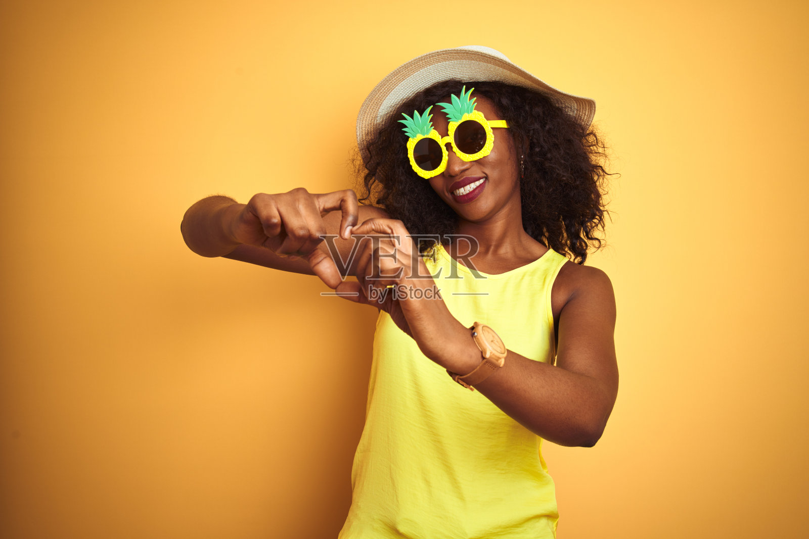 非洲裔美国妇女戴有趣的菠萝太阳镜在孤立的黄色背景微笑在爱显示心形符号和形状的手。浪漫的概念。照片摄影图片