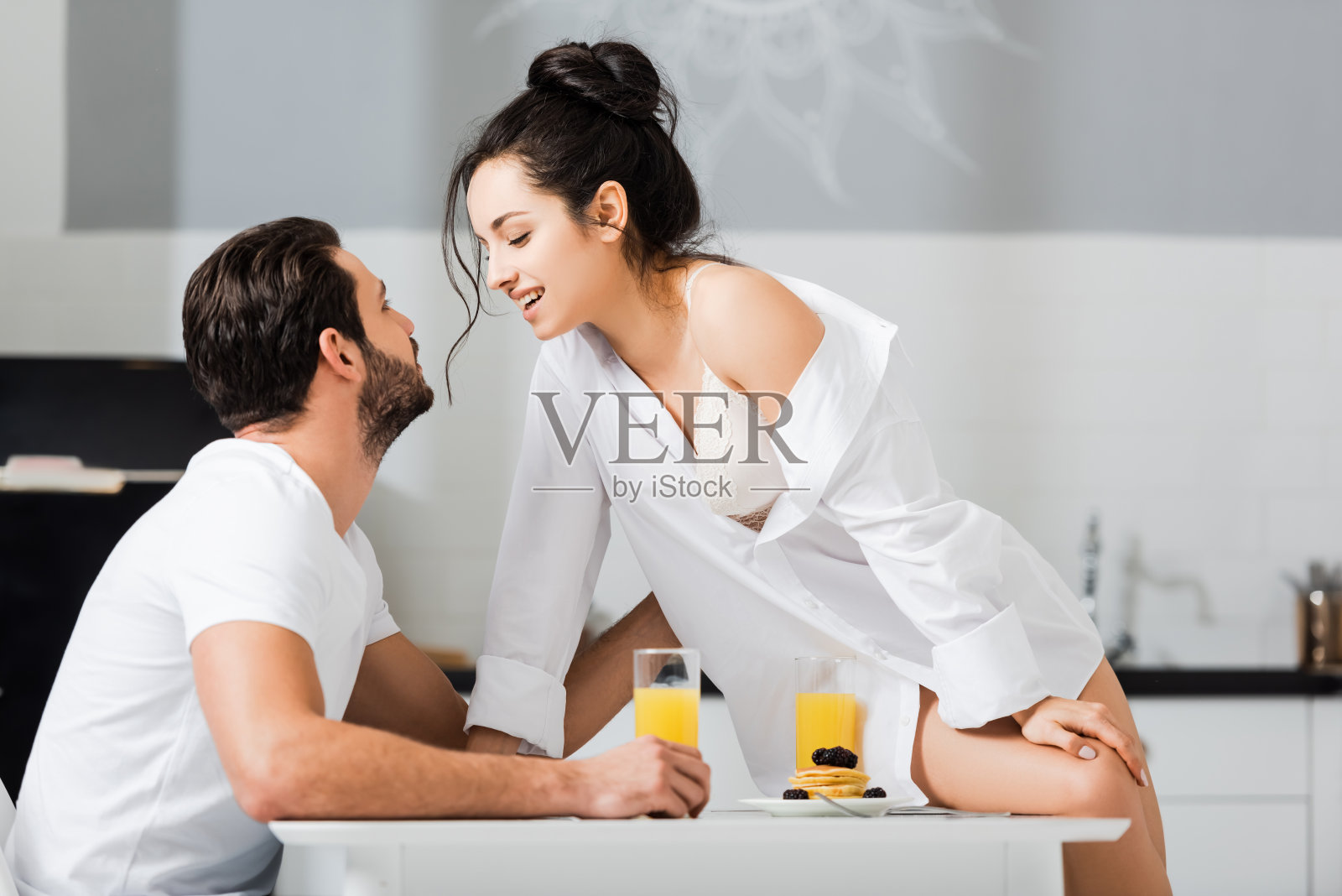 侧面的性感女人在衬衫和胸罩微笑的男朋友，而坐在厨房的桌子上照片摄影图片