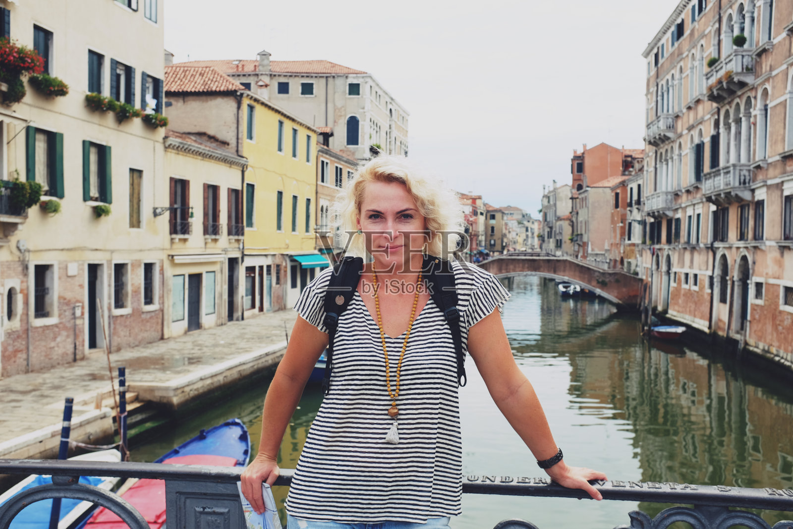 穿着条纹衬衫的金发美女站在威尼斯的一座步行桥上，微笑着看着镜头，背景是一条运河和老房子照片摄影图片