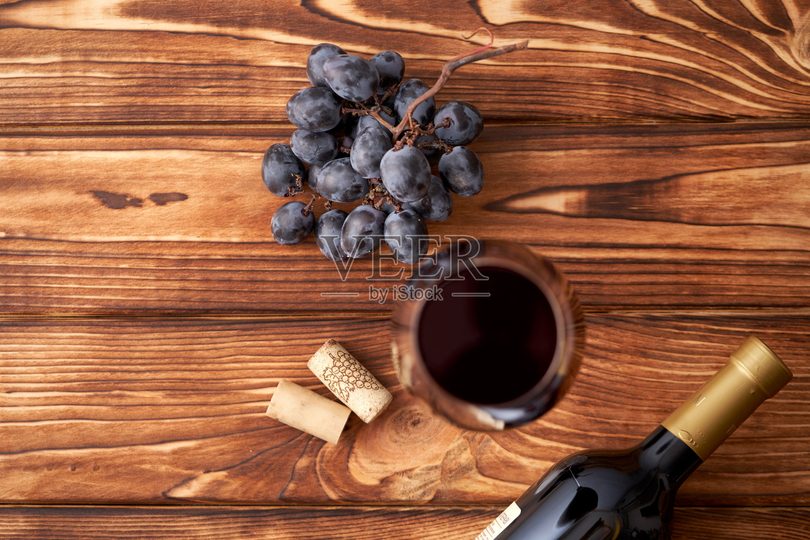 一串成熟的蓝葡萄，一杯红酒，旁边有酒塞，一瓶密封的红酒，背景是有纹理的木头。酿酒、品酒照片摄影图片