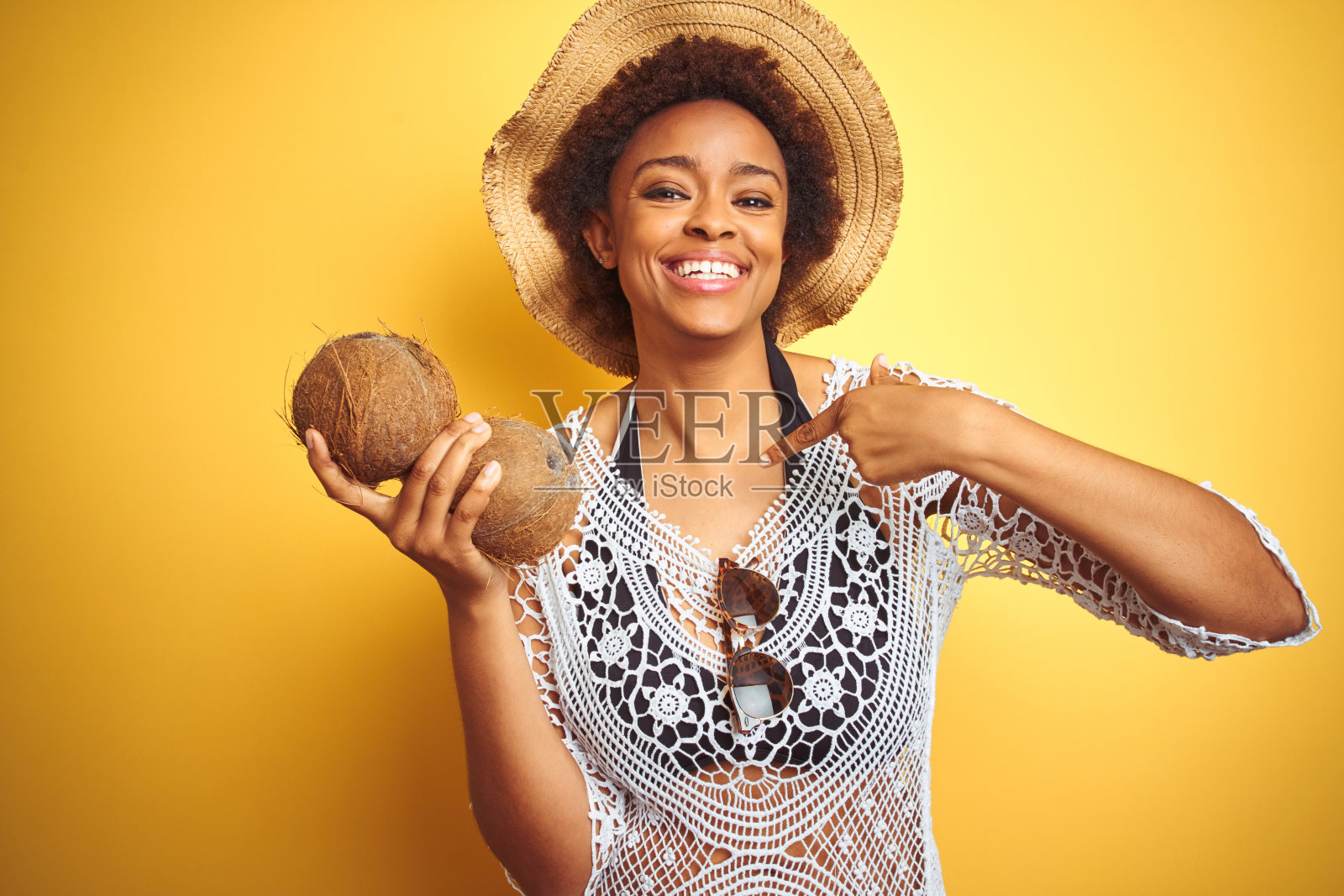 年轻的非洲裔美国妇女与非洲式的头发拿椰子在黄色孤立的背景与惊讶的脸指向自己照片摄影图片