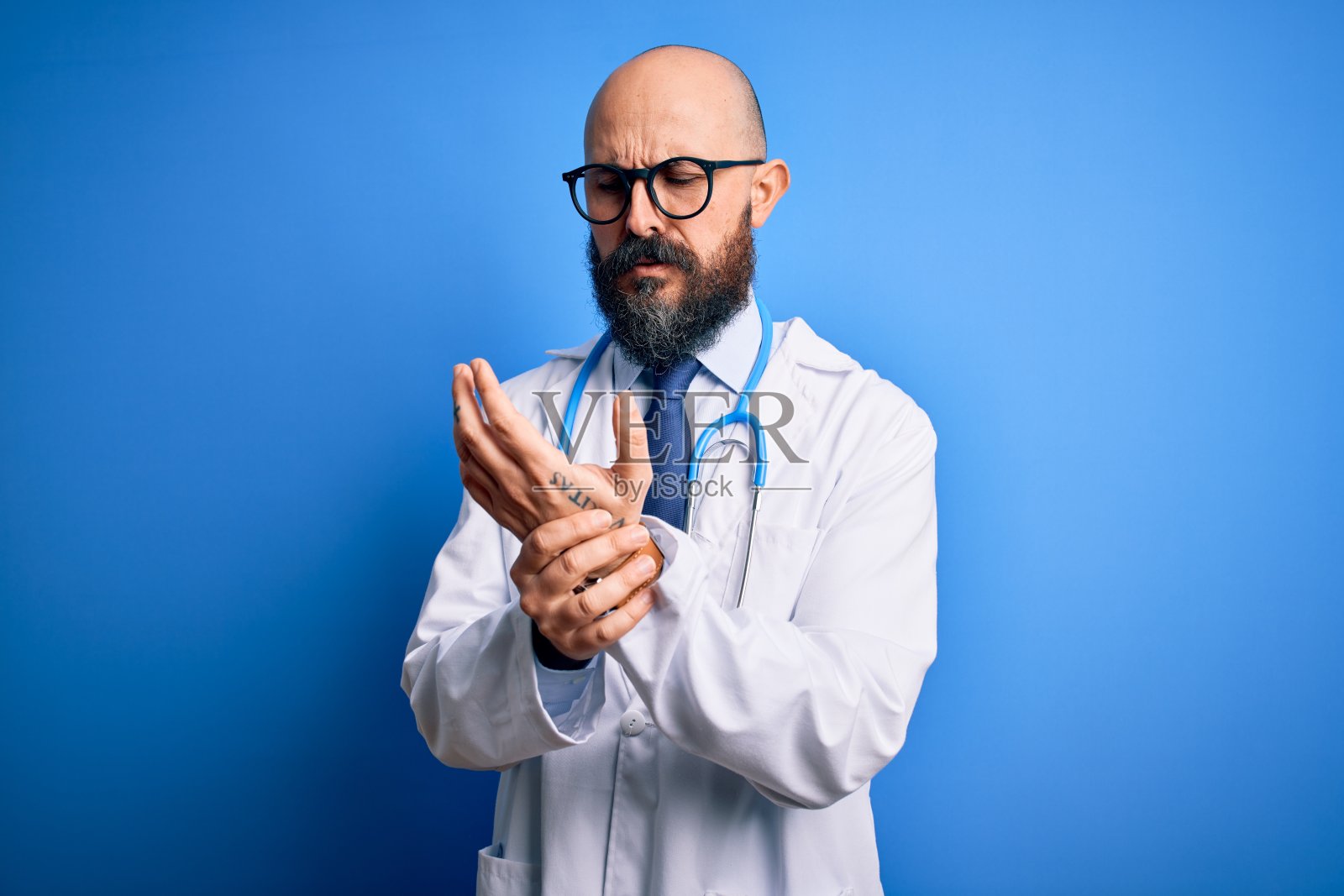 英俊秃顶的医生男子，留着胡子，戴着眼镜和听诊器，背景是蓝色。手和手指疼痛，关节炎发炎照片摄影图片