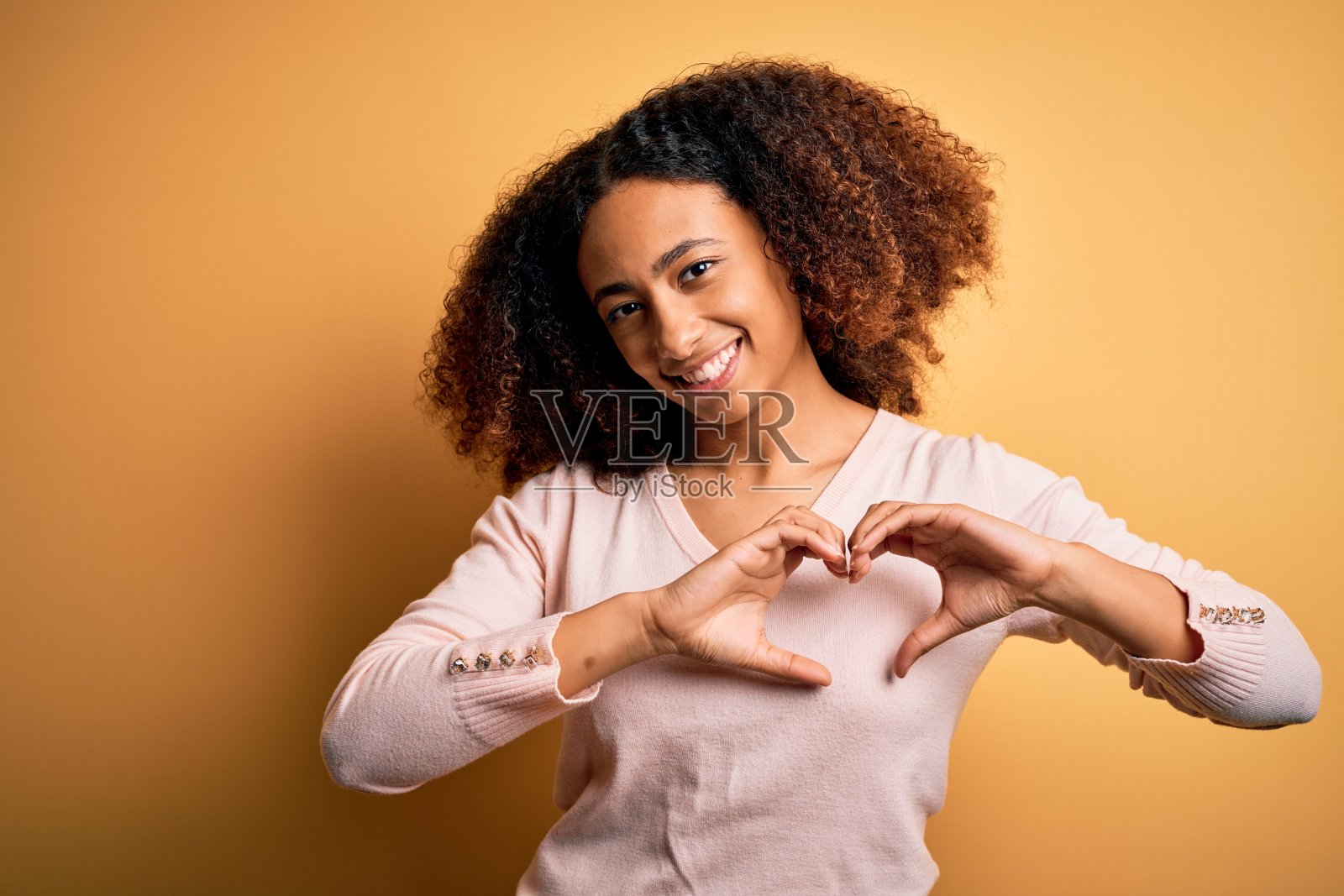 年轻的非洲裔美国妇女与非洲发式穿着休闲毛衣在黄色的背景微笑在爱显示心形符号和形状的手。浪漫的概念。照片摄影图片
