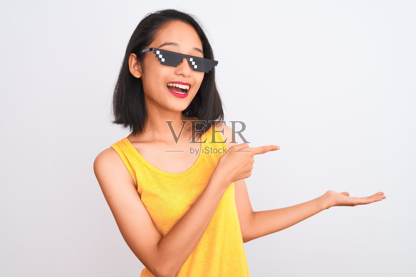 年轻美丽的中国女子戴着暴徒生活太阳镜，在孤立的白色背景下，对着镜头惊讶地微笑着，同时用手指着镜头。照片摄影图片