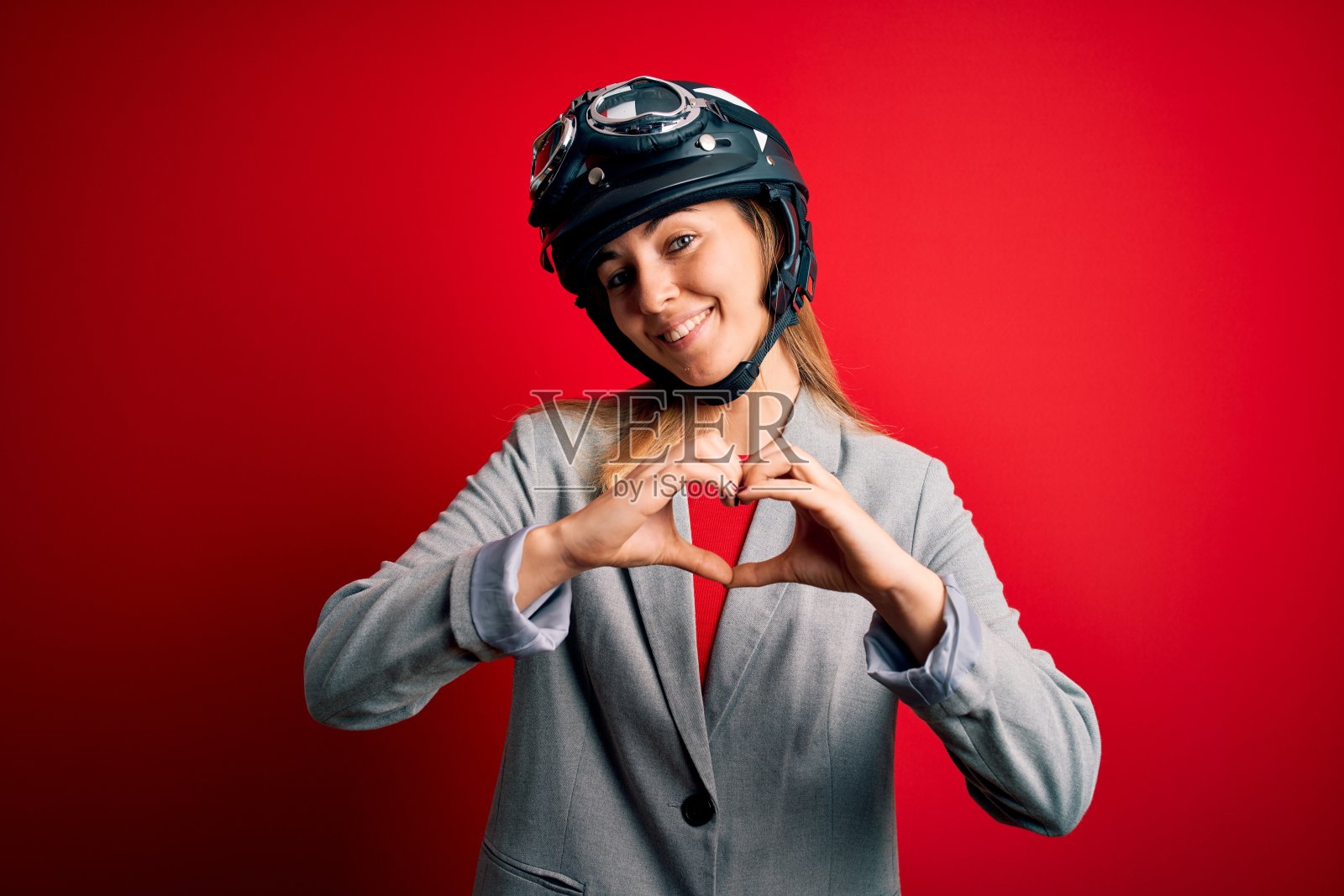 年轻美丽的金发摩托车女戴着摩托车头盔在红色的背景微笑着做心形符号的手。浪漫的概念。照片摄影图片