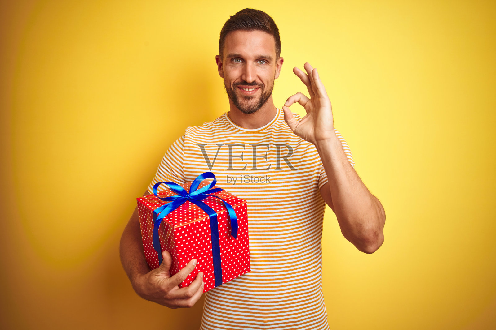 一个年轻人拿着一份生日礼物在黄色的孤立的背景下用手指做ok手势，很好的象征照片摄影图片