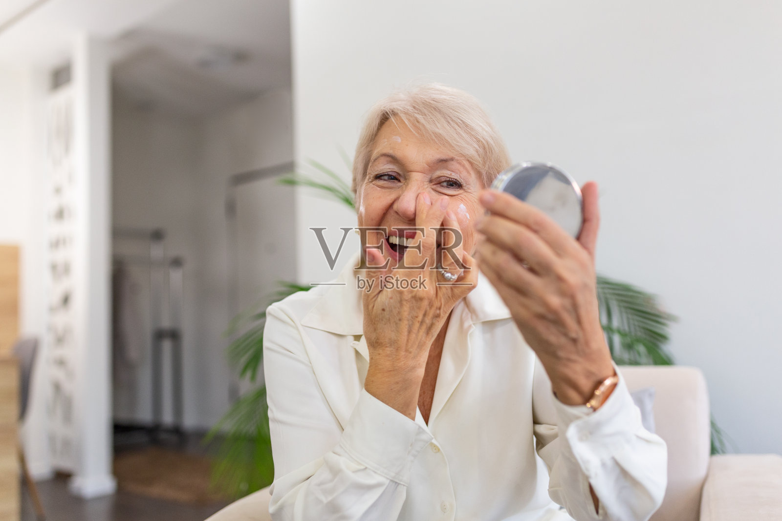 微笑的老年妇女使用抗衰老乳液，以消除黑眼圈。快乐成熟的女人用化妆霜来隐藏眼睛下面的皱纹。女士使用日霜。照片摄影图片