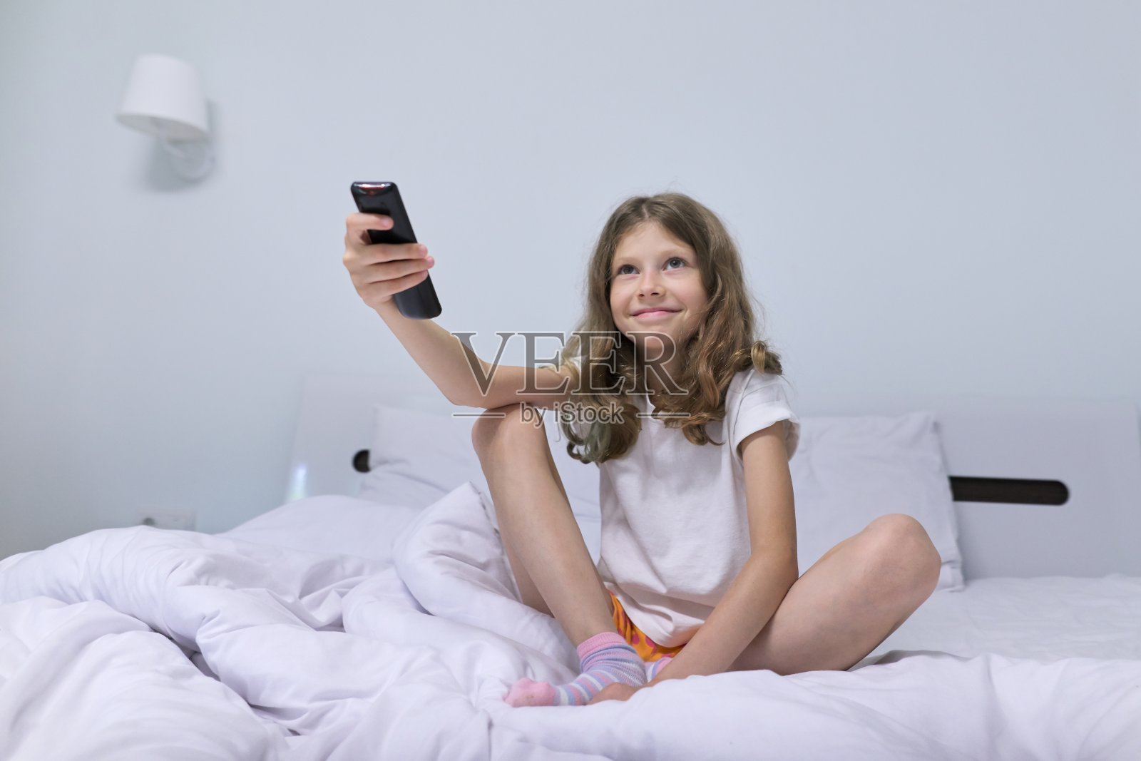 漂亮的金发女孩早上坐在白色的床上，拿着电视遥控器照片摄影图片