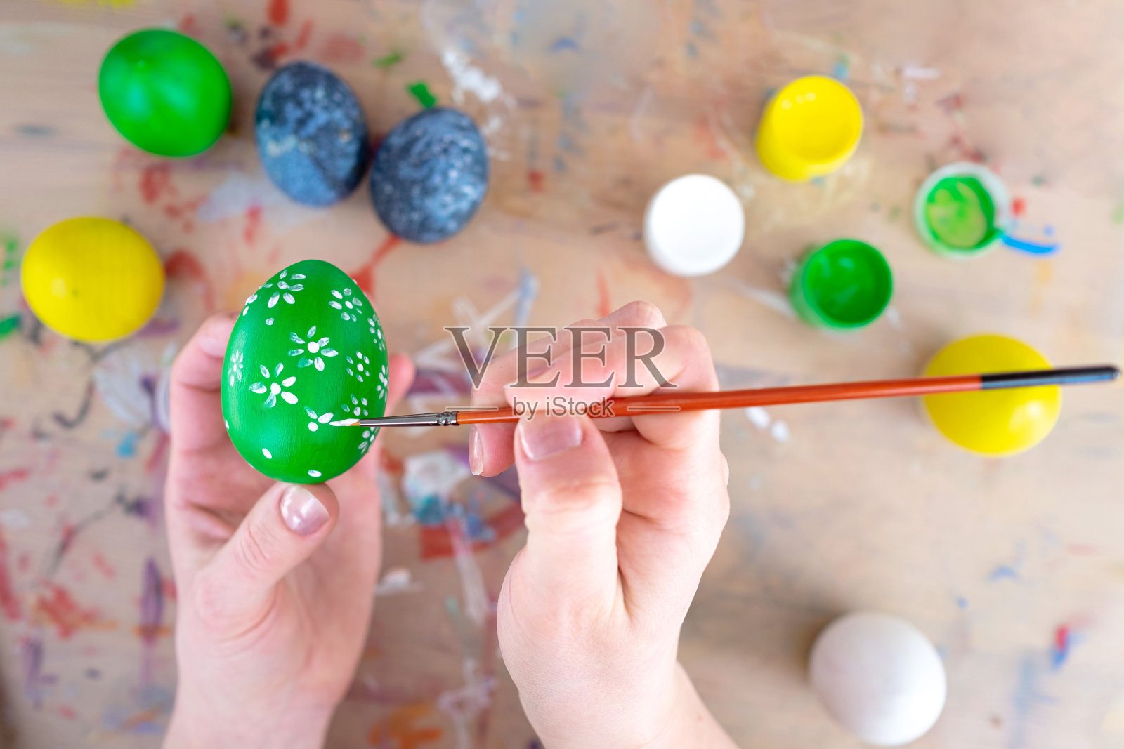 庆祝复活节的概念。女人的手在一个绿色的鸡蛋上画上花朵图案。有油漆罐和彩色彩蛋的工作场所照片摄影图片