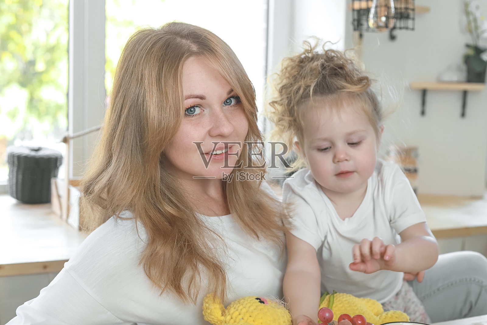 室内拍摄的漂亮的金发母亲和她的小女儿给予拥抱，享受家庭气氛，对着宽敞的白色厨房室内摆姿势。母亲和家庭观念。照片摄影图片