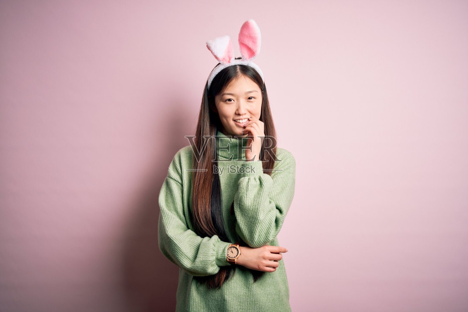 一名年轻的亚洲女子，在粉红色的背景下戴着可爱的复活节兔子耳朵，看起来很自信地看着镜头，双臂交叉，手托着下巴。积极的思考。照片摄影图片