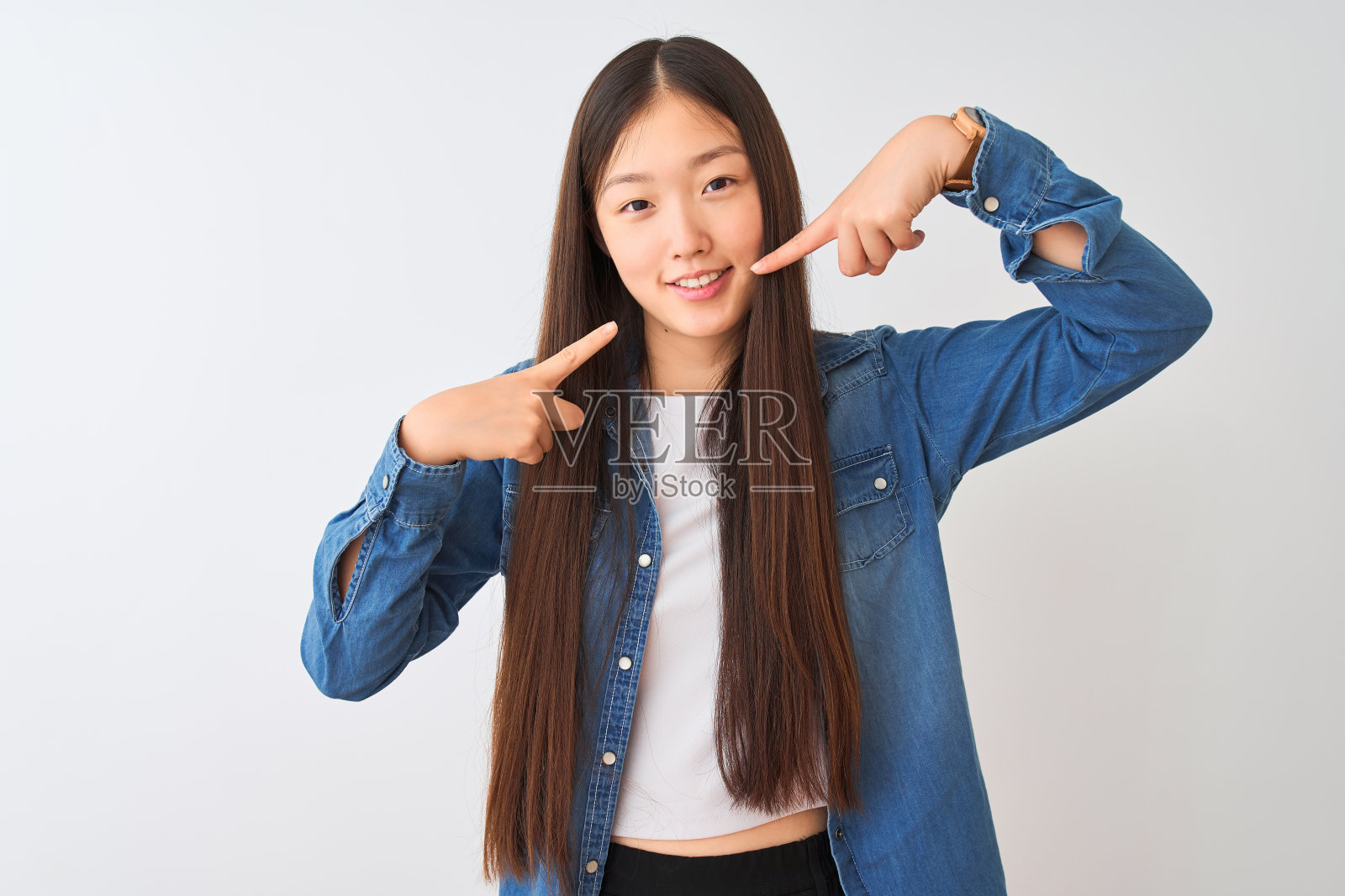 年轻的中国妇女穿着牛仔衬衫站在孤立的白色背景微笑，显示和指向手指，牙齿和嘴。牙齿健康的概念。照片摄影图片