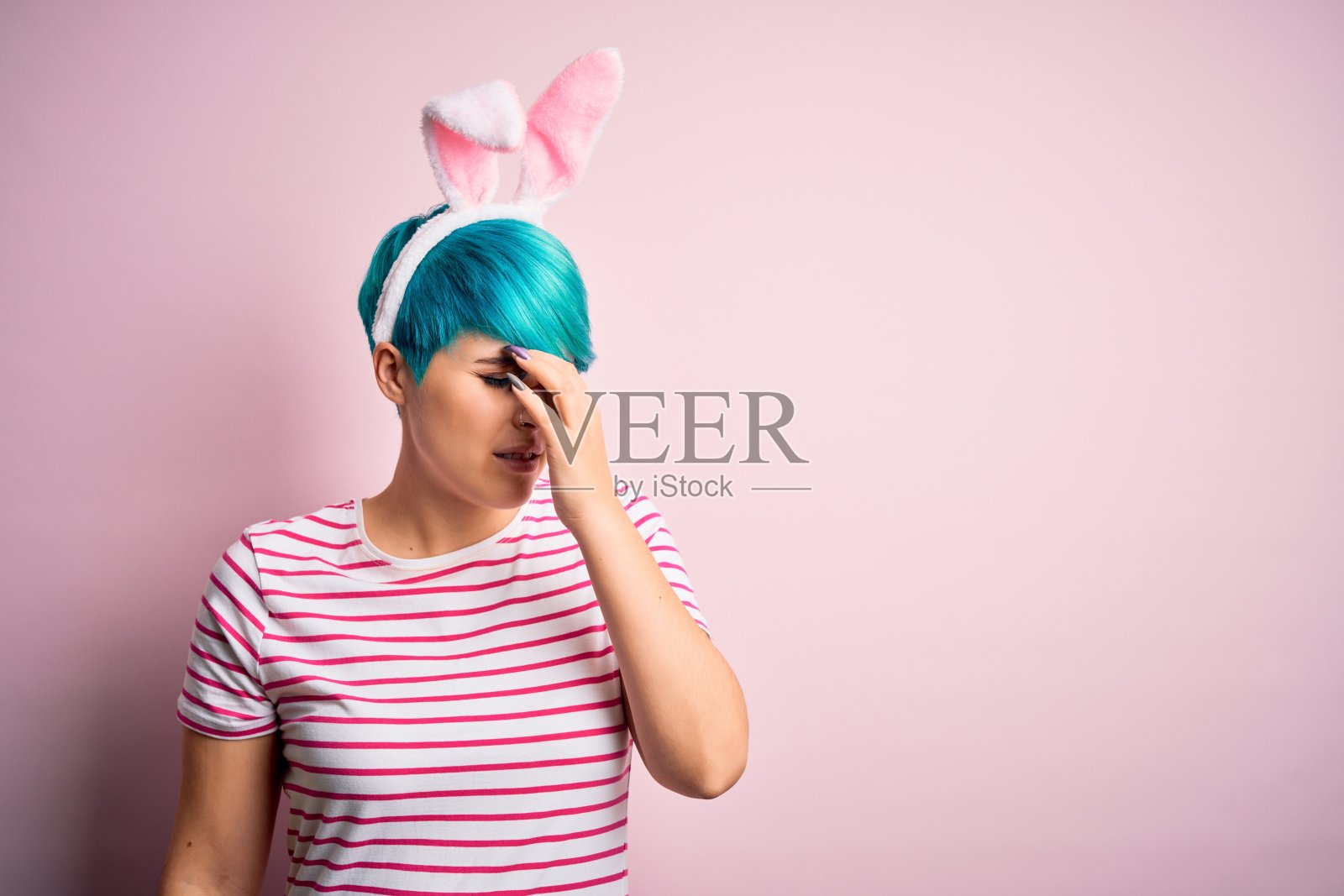 年轻女子与时尚的蓝色头发戴复活节兔子耳朵在粉红色的背景疲劳摩擦鼻子和眼睛感到疲劳和头痛。压力和挫折的概念。照片摄影图片