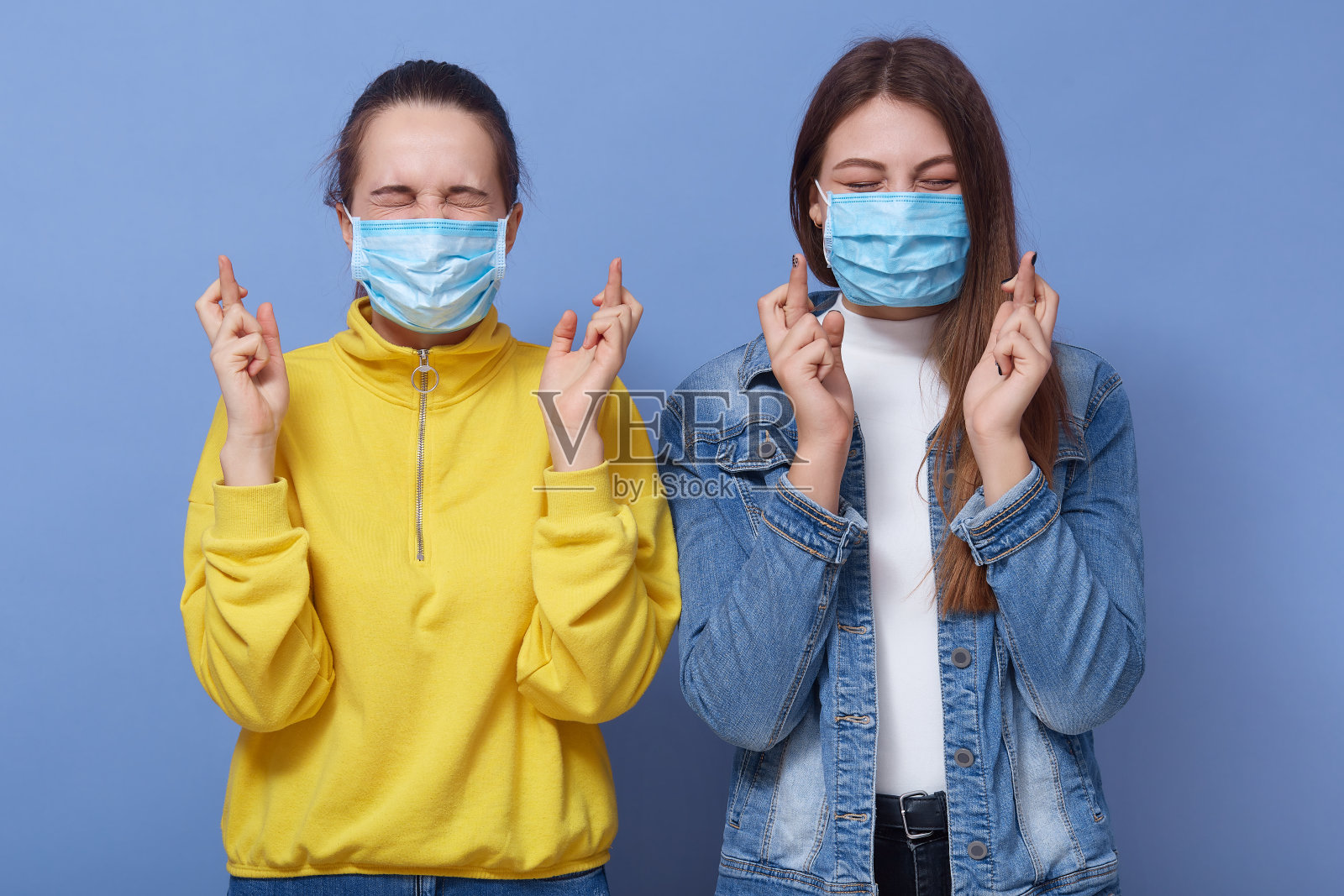 近距离肖像的两个年轻妇女交叉手指，戴防护口罩，防止病毒感染，女性穿着休闲服装，摆孤立的蓝色背景。Covid 19的概念。照片摄影图片