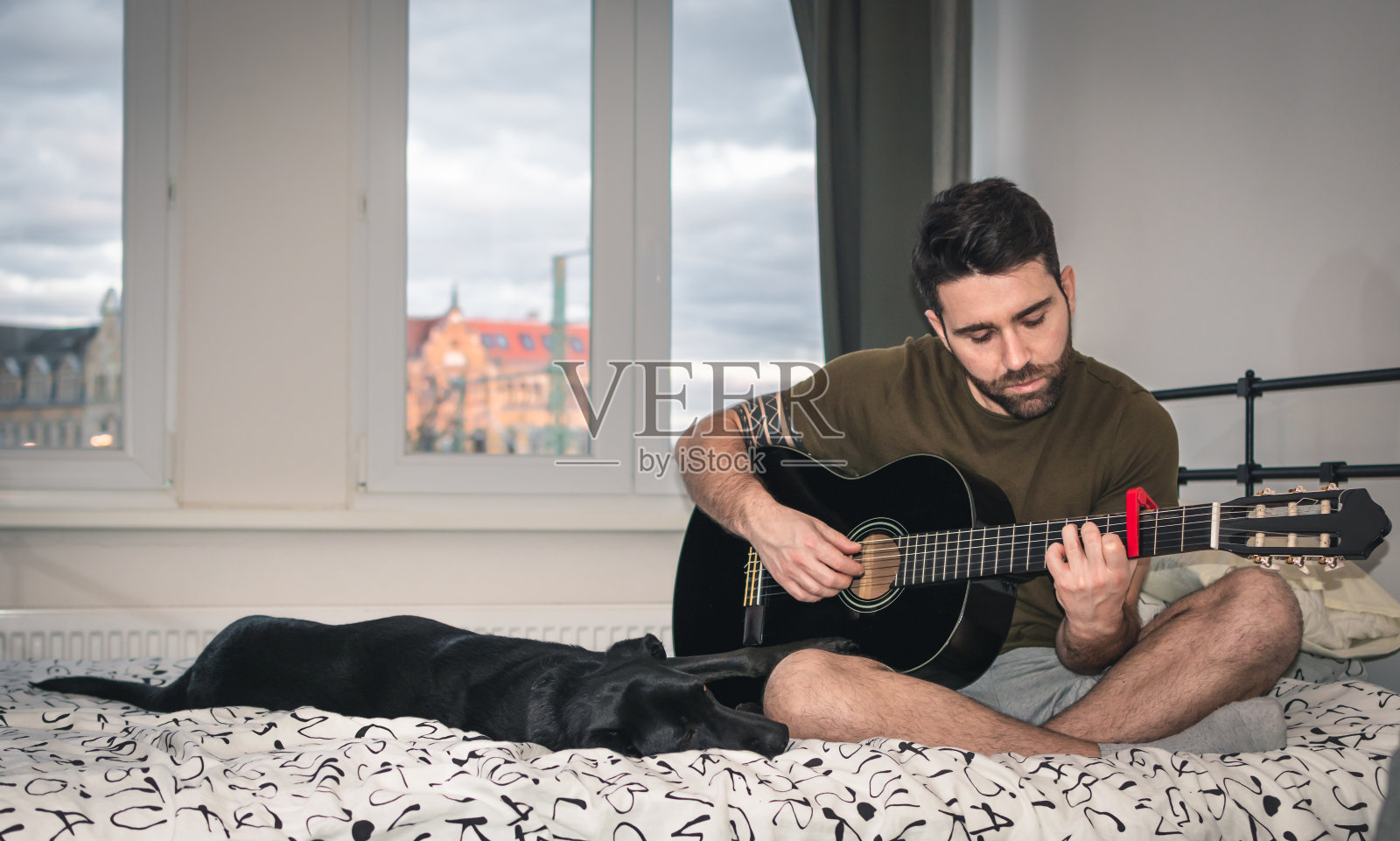 男孩独自在家，带着悲伤的表情，弹着吉他，坐在床上，旁边是他的狗，前面是一扇明亮的窗户，在那里你可以看到城市照片摄影图片