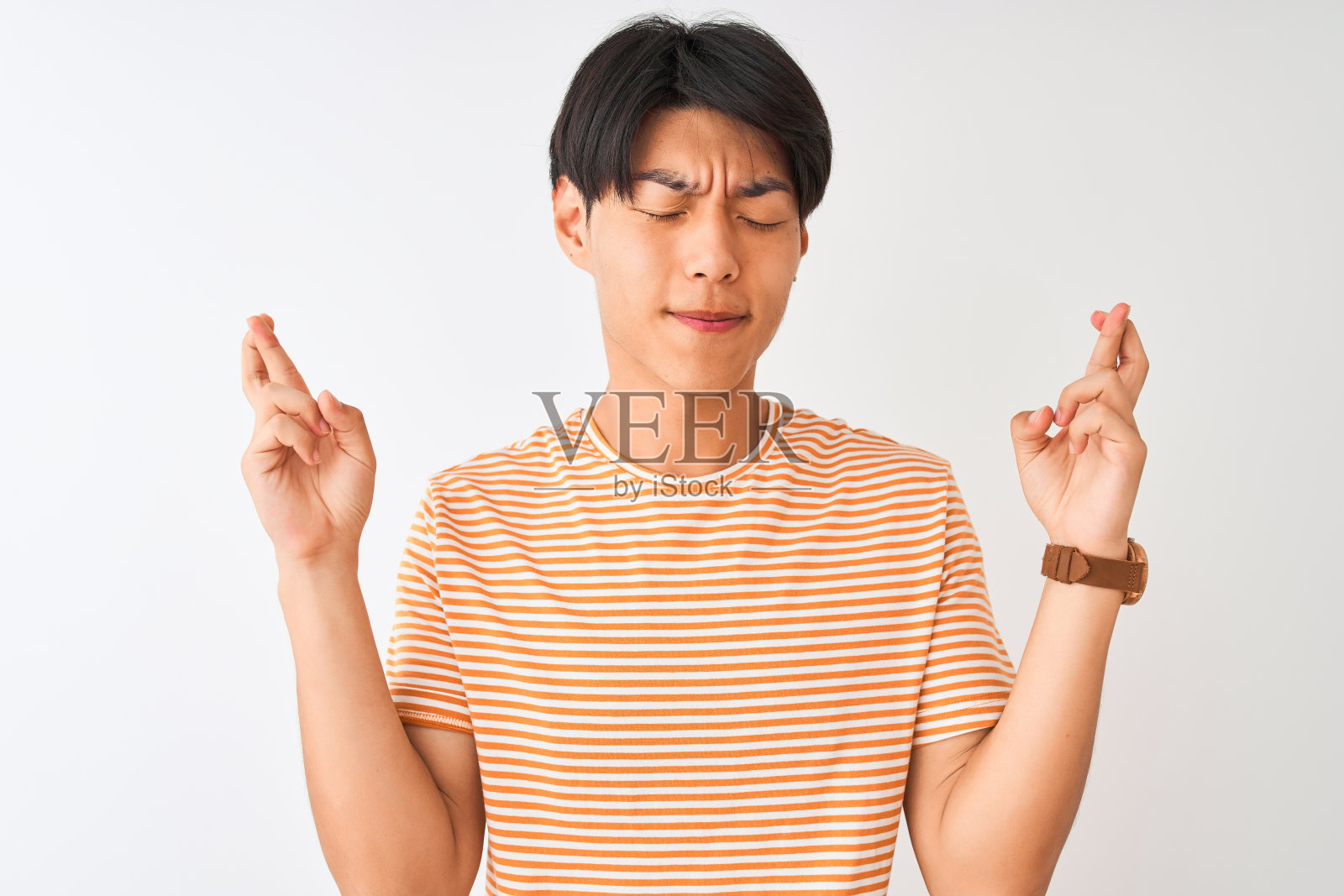 年轻的中国男子穿着休闲条纹t恤，站在孤立的白色背景下，双手交叉手指，闭上眼睛，满怀希望地微笑。运气和迷信的概念。照片摄影图片