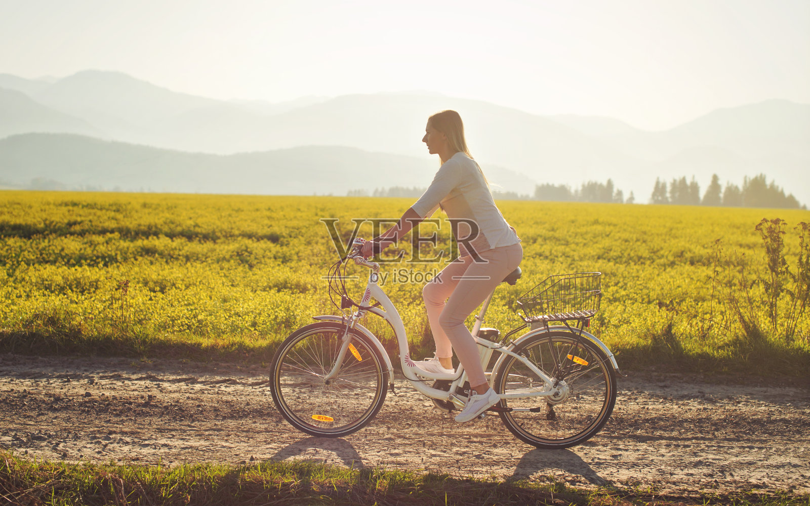 一名年轻女子骑着电动车在尘土飞扬的乡村道路上行驶，午后强烈的太阳背光在背景中照射在黄花田上，从侧面观看照片摄影图片