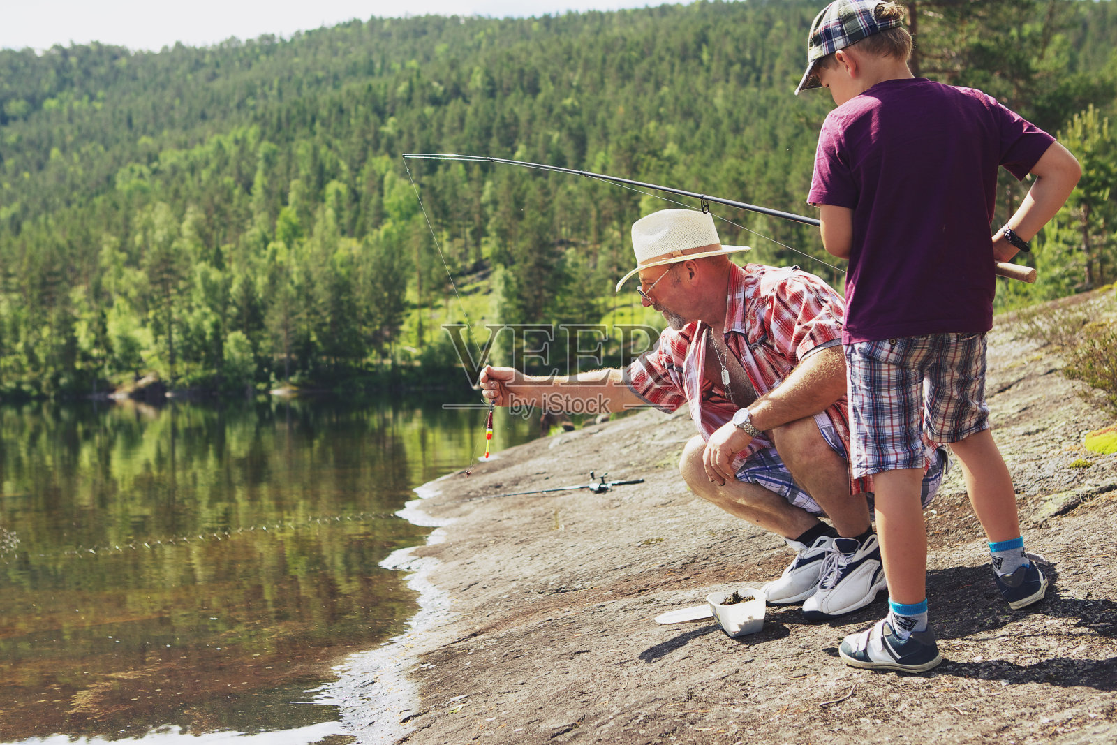 一个时尚的爷爷和孙子在一起——爷爷正在帮助小男孩拿着他的鱼竿，站在挪威山区湖边的一块阳光下的岩石上照片摄影图片