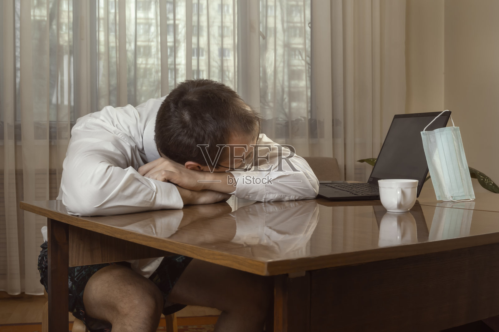 一名身穿白衬衫、打着领带、穿着内衣的年轻人疲惫地在家里的电脑前工作，把头靠在桌子上。照片摄影图片