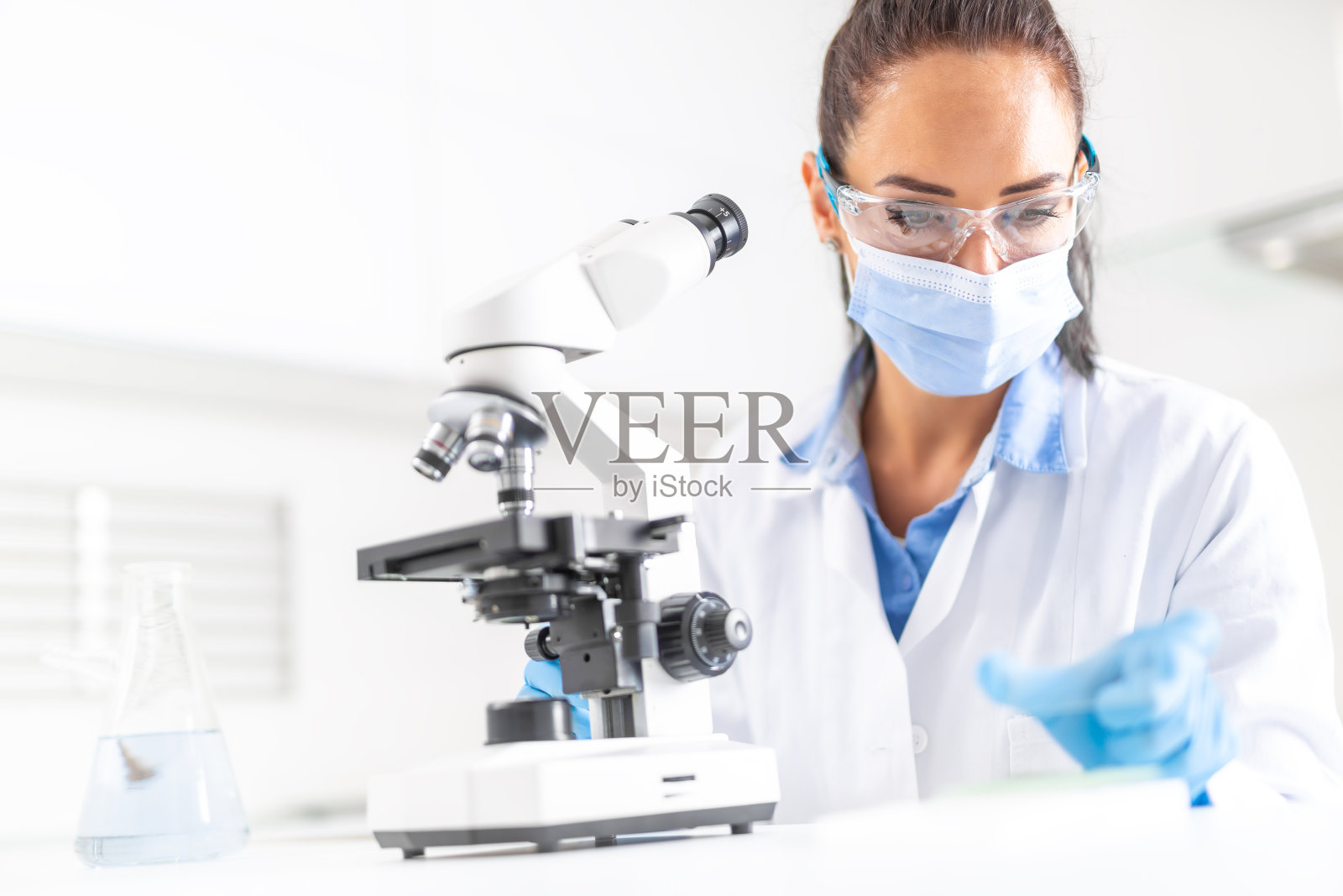 戴着防护眼镜、手套和面罩的女性实验室技术员坐在显微镜和锥形瓶旁边，看着桌子旁边。照片摄影图片