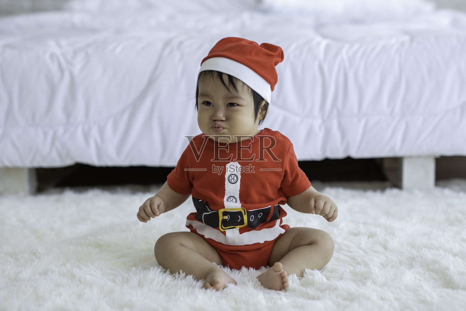 可爱可爱的亚洲宝宝戴着圣诞老人的帽子和布料服装坐在白色的地毯上微笑，在舒适的卧室玩幸福的情绪，健康的宝宝概念照片摄影图片
