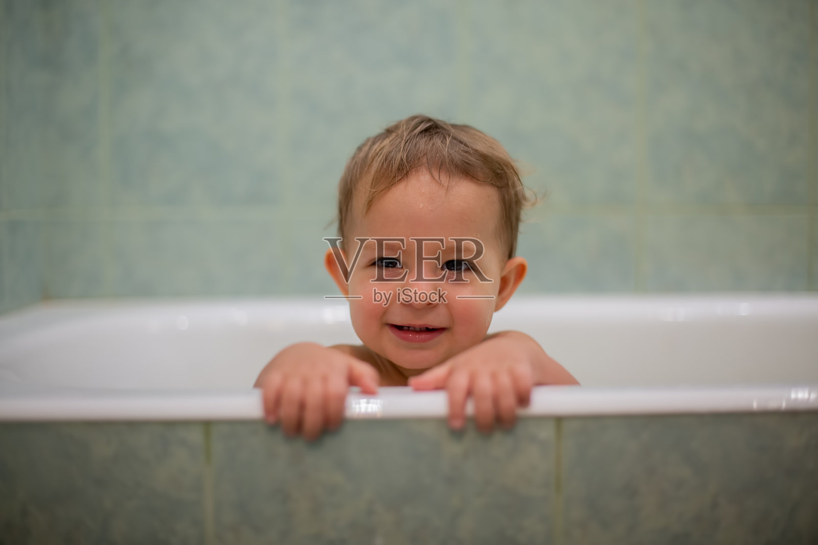 一个可爱的白人婴儿从浴缸里探出头来，把手放在浴缸边，笑着看着镜头。背景是一个模糊的绿色浴室。特写,软焦点照片摄影图片
