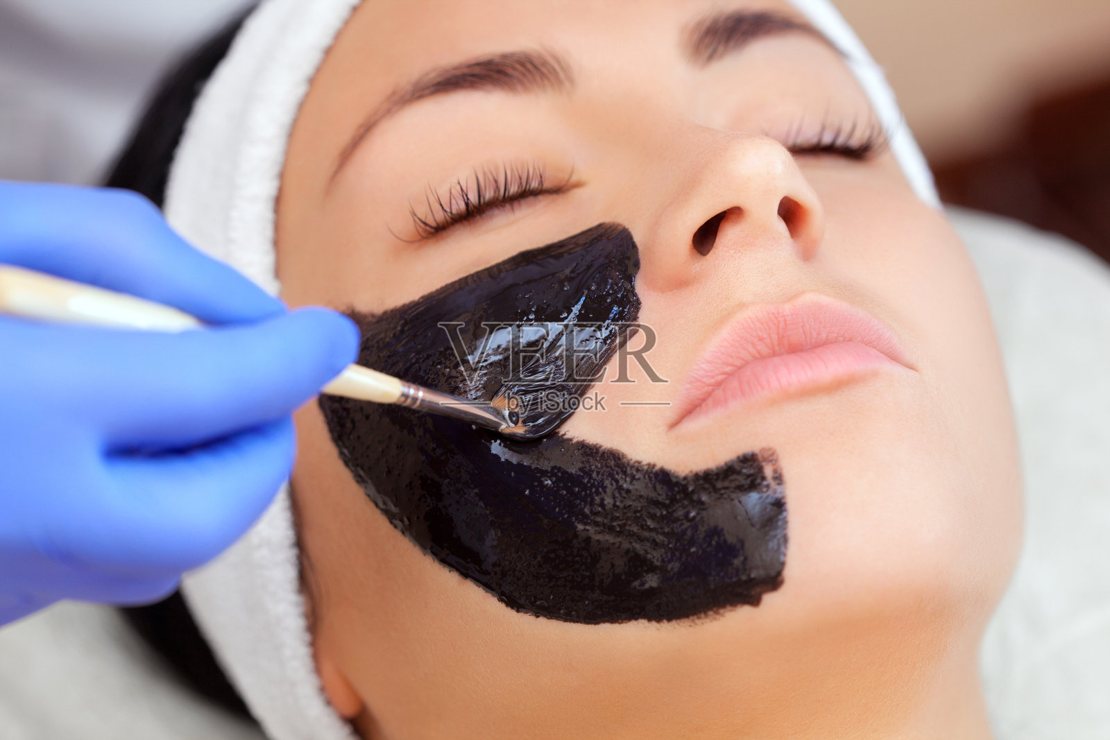 美容师做一个黑色面膜清洁皮肤和抗皱在一个美丽的女人的脸上，水疗和美容的概念。照片摄影图片