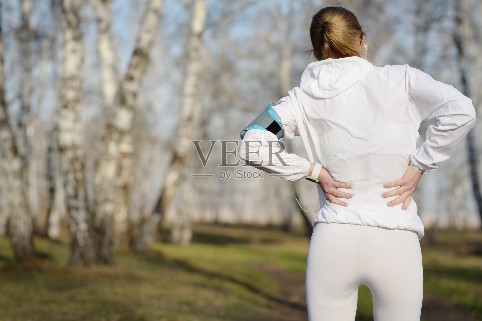 背部疼痛。运动妇女在白色运动服站在公园摩擦她的下背部肌肉。照片摄影图片