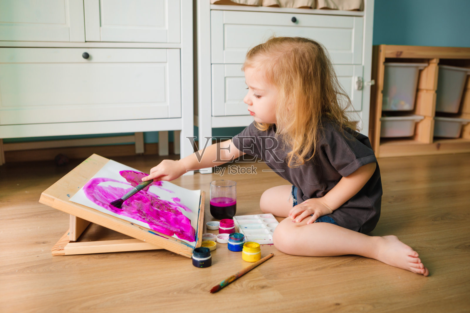 一个孩子坐在儿童房间的地板上，用颜料在画架上画画。照片摄影图片