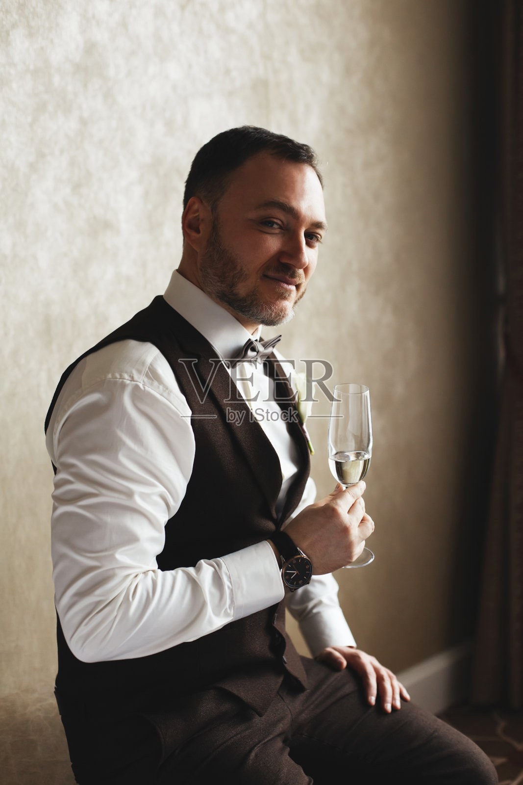 一个英俊的新郎在酒店房间里拿着一杯香槟的肖像。照片摄影图片