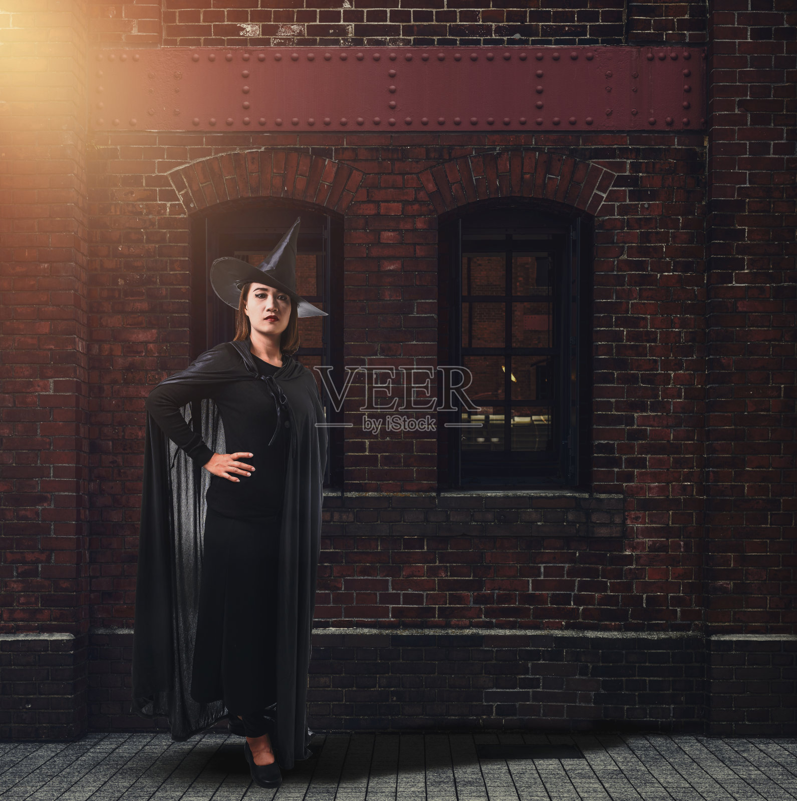 女人在黑色可怕的巫婆万圣节服装与老砖墙的古老城堡的女巫照片摄影图片