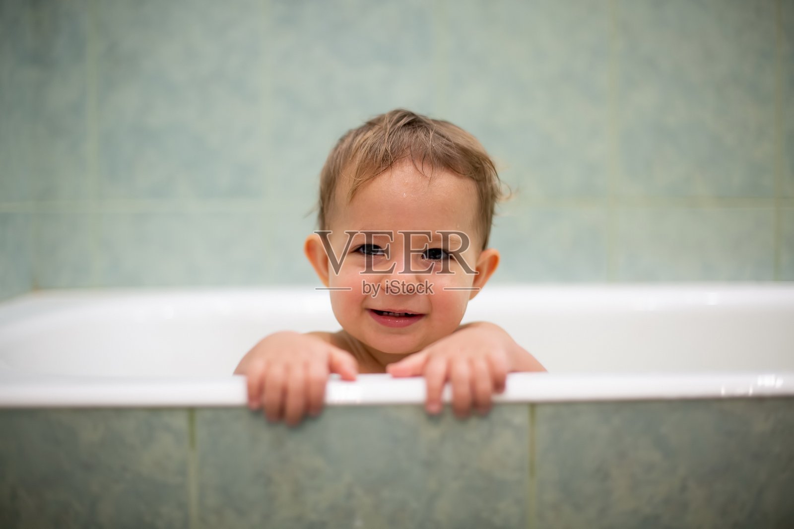 一个可爱的白人婴儿从浴缸里探出头来，把手放在浴缸边，笑着看着镜头。背景是一个模糊的绿色浴室。特写,软焦点照片摄影图片