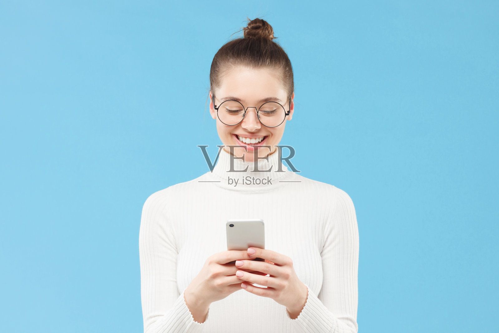 年轻的女学生穿着白色毛衣，戴着眼镜，手里拿着手机，与朋友们交换短信，孤立在蓝色背景下照片摄影图片