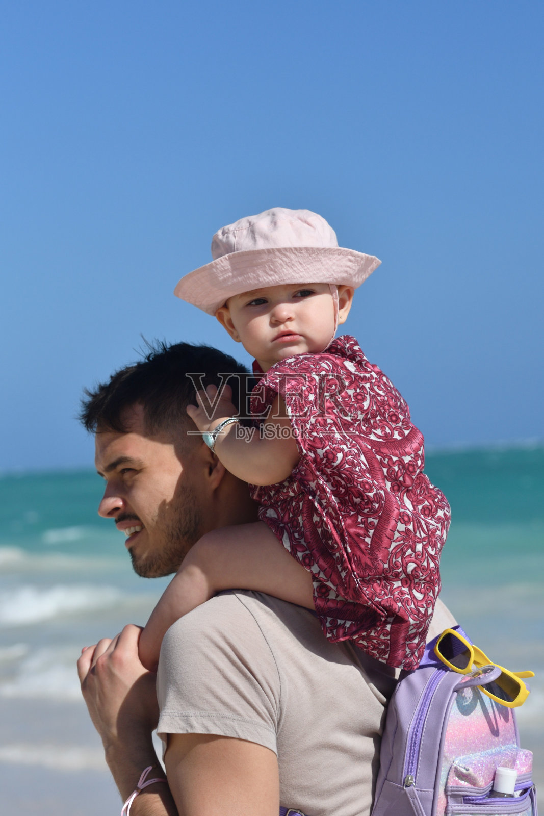 在阳光明媚的热带海滩，父亲背着15个月大的蹒跚学步的孩子照片摄影图片
