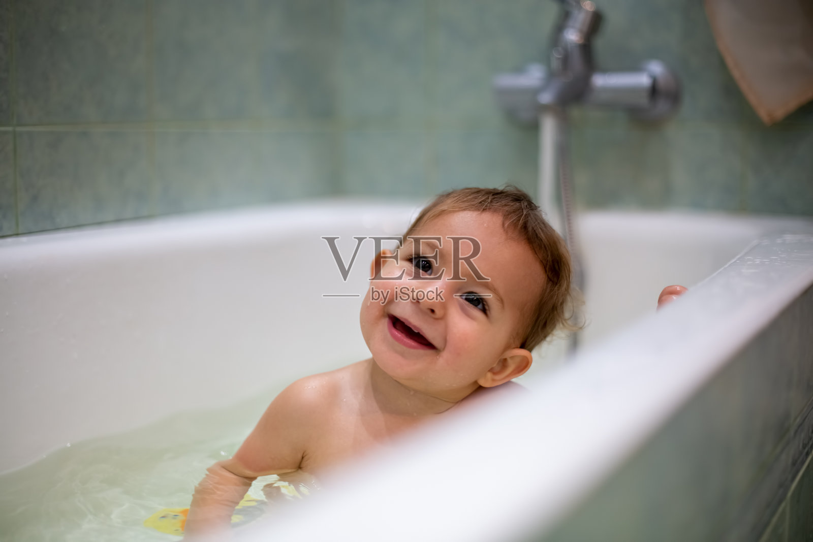 可爱的白人婴儿正在洗澡，微笑着抬头看，水花飞溅，背景是一间模糊的绿色浴室。特写,软焦点照片摄影图片