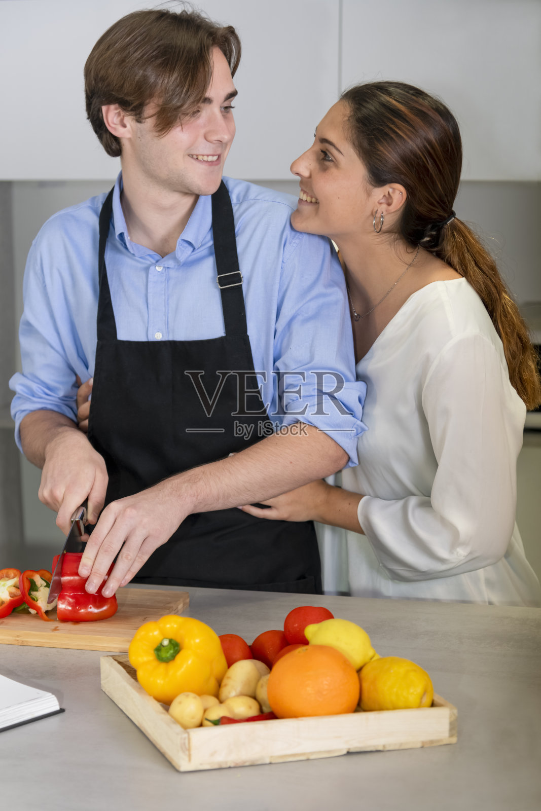 一对年轻的夫妇看着对方，在一个厨房里，在一个模糊的背景上切一个甜椒。照片摄影图片