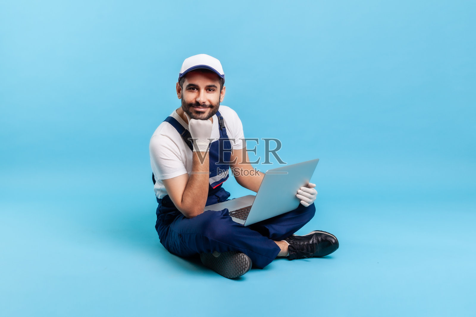 一个快乐的职业杂工的肖像，穿着工作服，盘腿坐在电脑上，靠在手上照片摄影图片