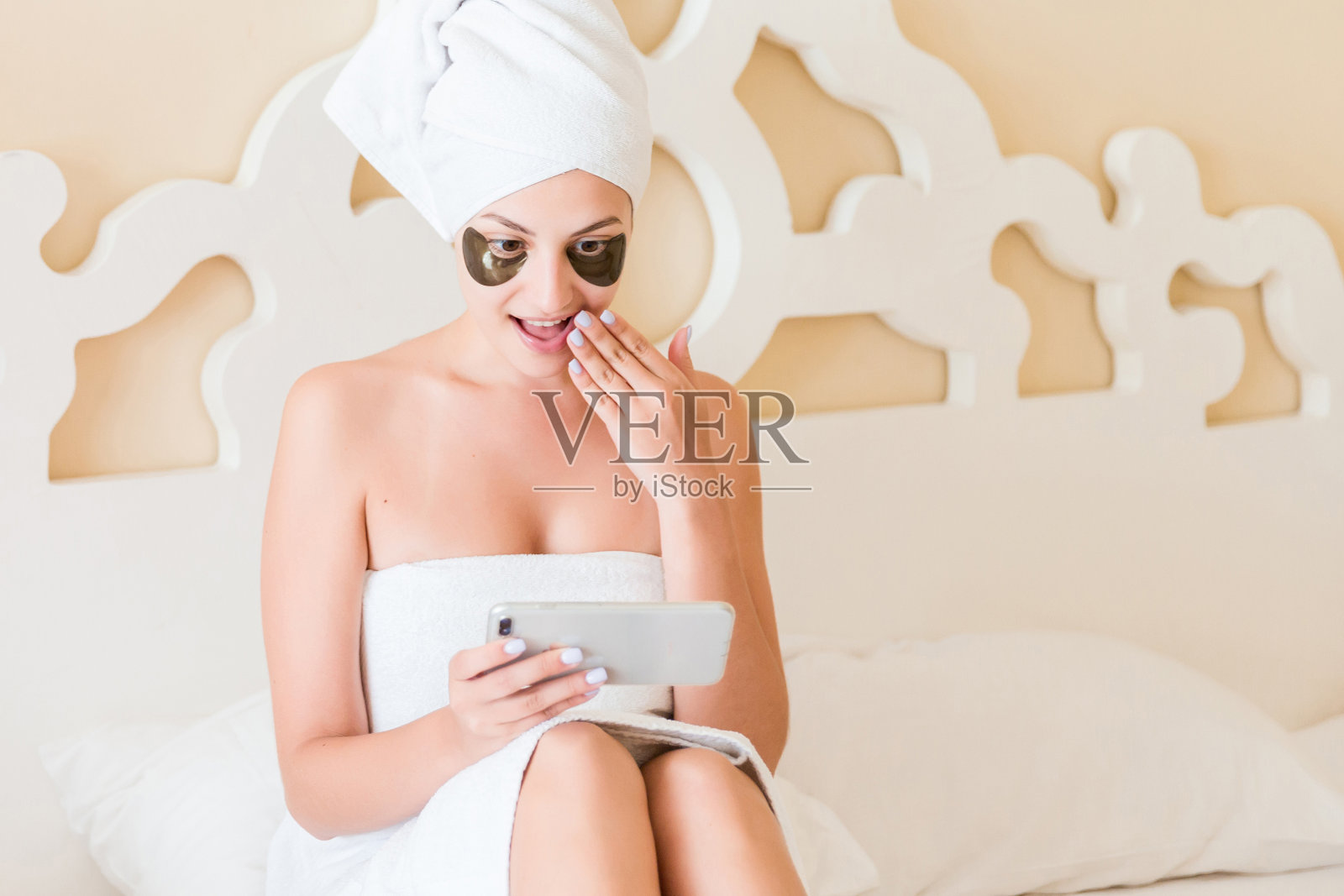 一个惊讶的女人在早上淋浴后裹着毛巾，眼睛下面有黑色的眼罩，正在床上放松和使用她的手机。消除黑眼圈。护肤概念照片摄影图片