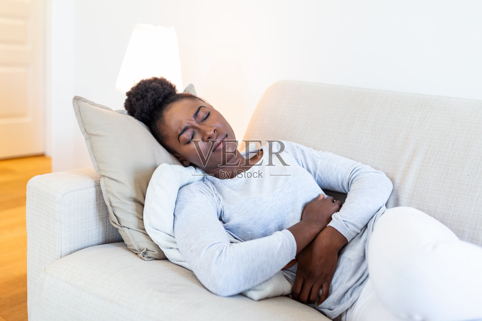 非洲裔美国妇女在痛苦的表情握着手对肚子遭受经期疼痛，躺在家里的床上伤心，有肚子抽筋的女性健康观念照片摄影图片
