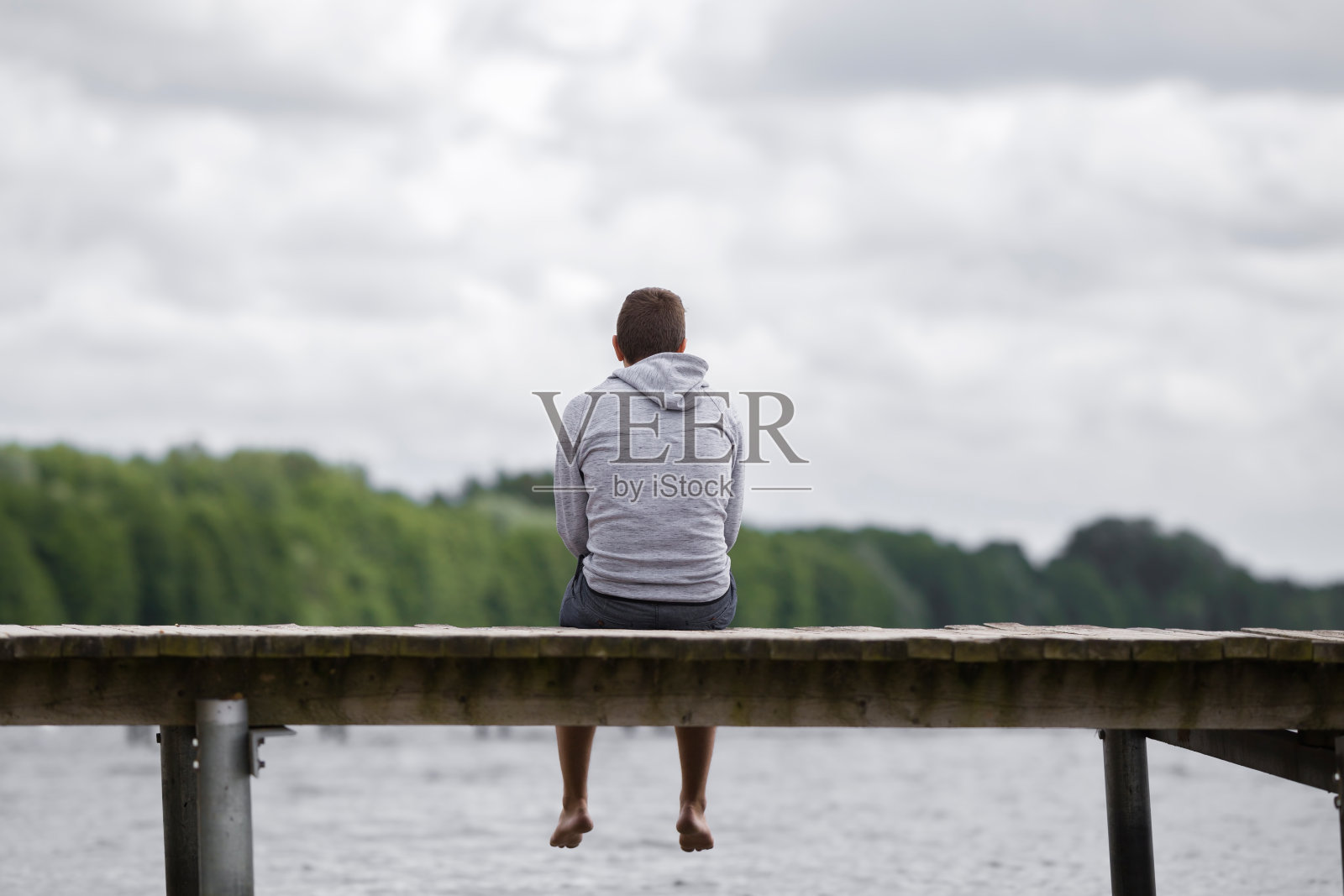 夏日，一个年轻人独自坐在人行桥边，凝视着湖水和多云的天空。思考人生。花时间独处在大自然中。和平的气氛。后视图。照片摄影图片