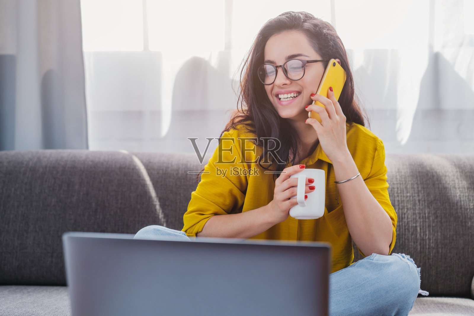 一个快乐的女孩，一边打电话，一边喝咖啡，一边坐在沙发上用笔记本电脑照片摄影图片