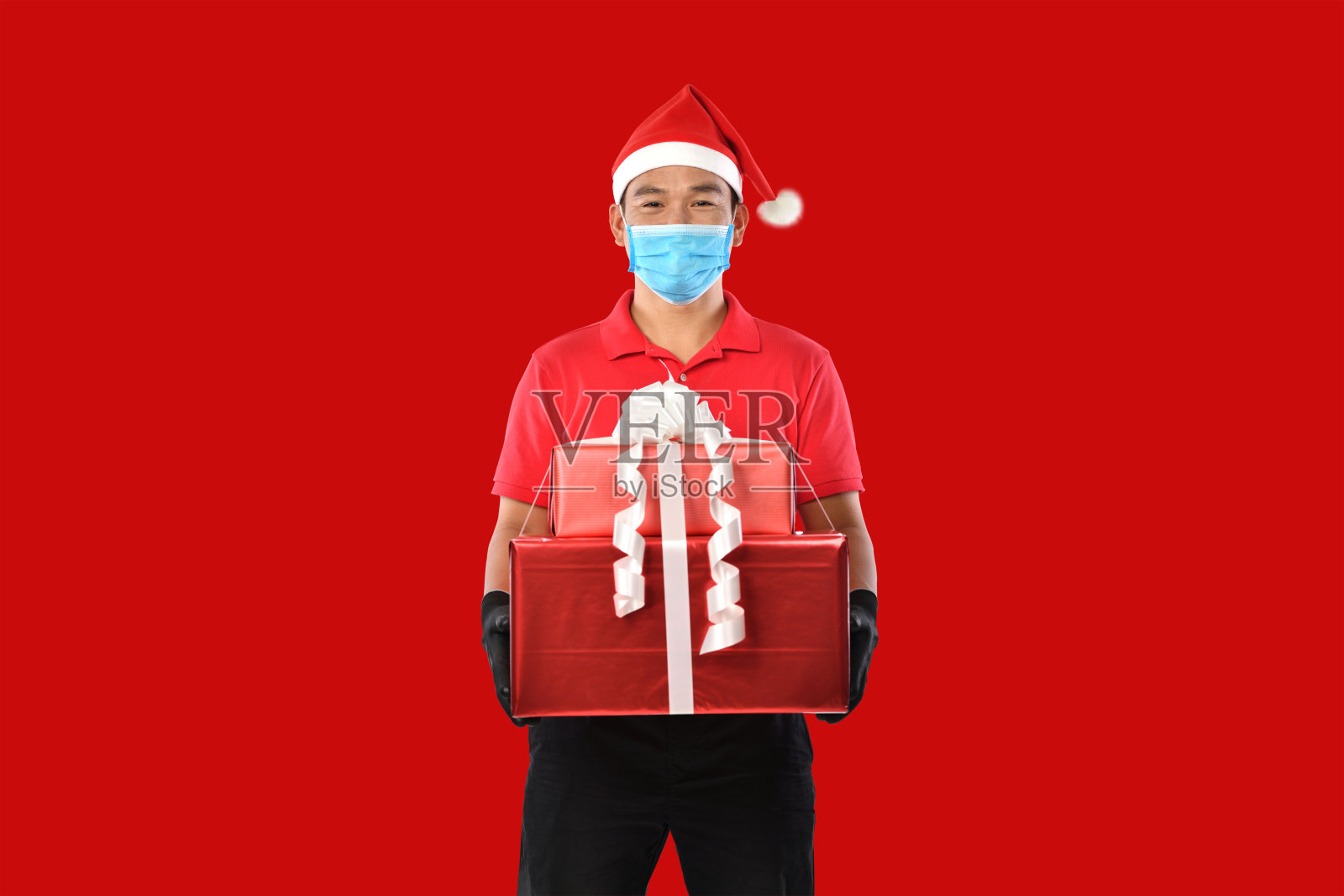 在新冠肺炎疫情爆发和圣诞节庆祝活动期间，身穿红色制服、医用口罩、防护手套、圣诞帽的快乐年轻亚洲快递员手持装有礼物的盒子照片摄影图片
