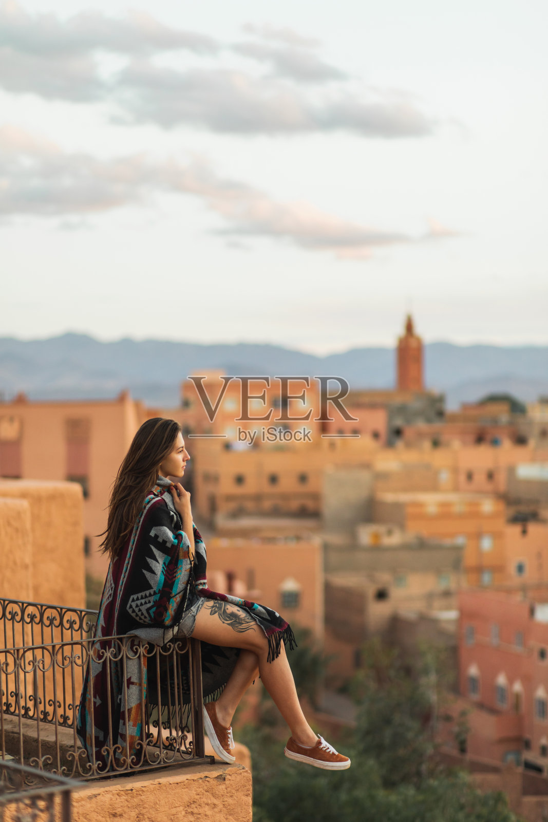 老妇人坐在屋顶露台上鸟瞰阿拉伯古镇布马恩。覆盖着传统的摩洛哥斗篷。嬉皮风格，裸腿纹身。照片摄影图片