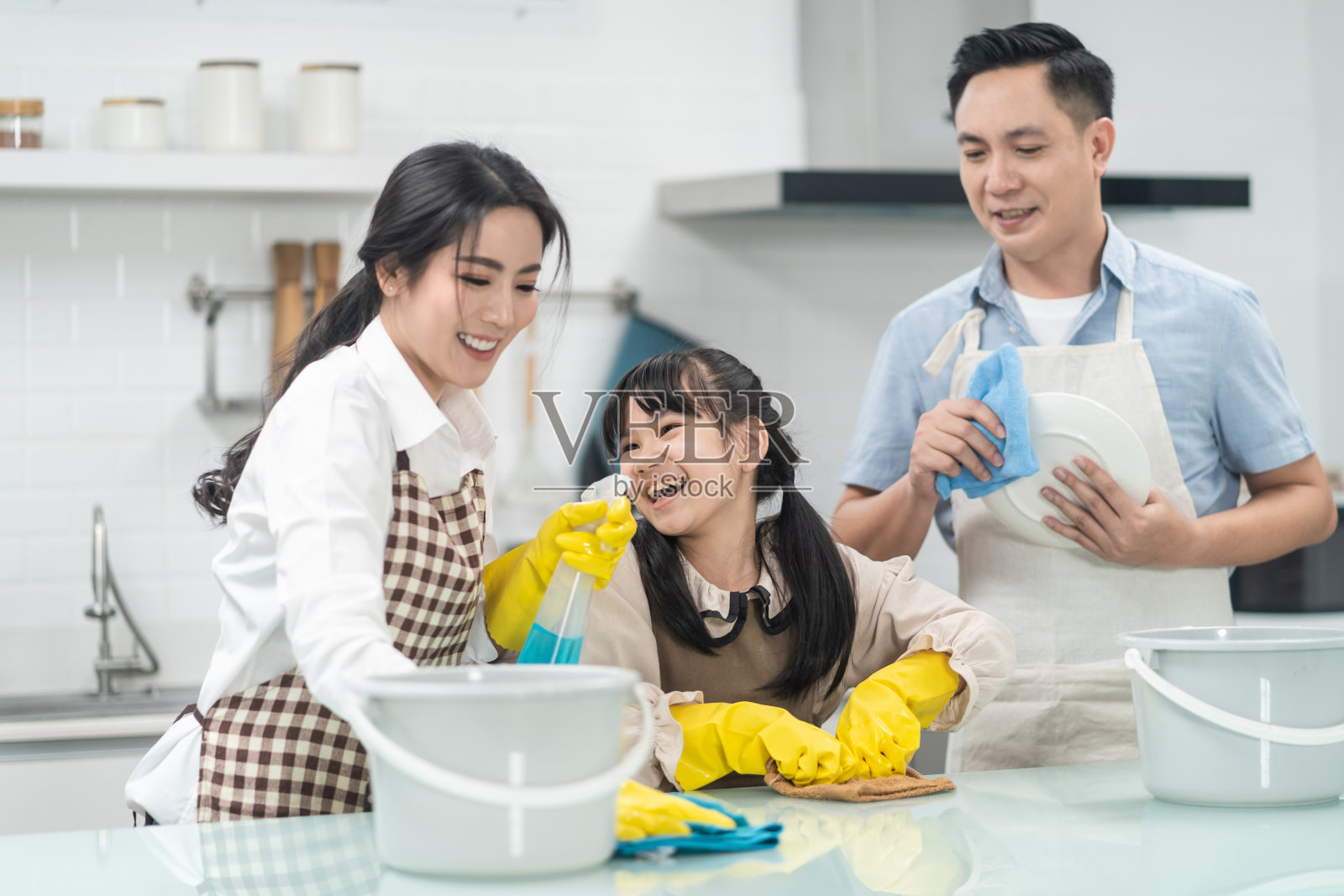 亚洲年轻家庭教他们的女儿清洁厨房柜台。孩子和父母一起跳舞，一起笑，很开心。可爱的家庭在快乐的打扫房间时的快乐时刻。照片摄影图片